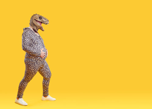 Funny fat man with big belly wearing pajamas and dinosaur mask isolated on orange background. Full length chubby man with dinosaur head and leopard pajamas stroking his belly near copy space.