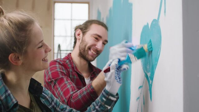 Smiling large charismatic couple enjoy the moment at home while painting the walls they make a big heart in a blue colour with small brushes and hugging each other with love
