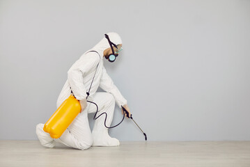 Pest control guy cleaning home from insects. Man in white protective suit crouching near wall and...