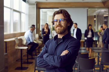 Portrait of happy male business teacher and professional coach. Handsome long haired bearded man in blue shirt and eyeglasses standing in office after corporate training class for team of employees - obrazy, fototapety, plakaty