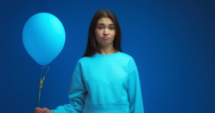 An upset oriental-looking woman wearing a blue jumper shows a sad and unhappy face because of one balloon on an isolated blue background