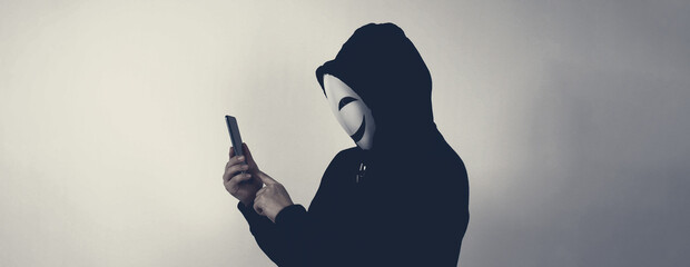 Anonymous hacker and face mask with smartphone in hand. Man in black hood shirt holding and using...