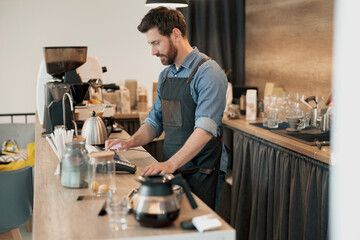 Fototapeta na wymiar Handsome barista wearing apron uses a digital tablet to takes order from clients at coffeehouse