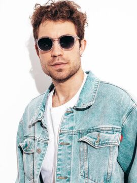 Portrait of handsome confident stylish hipster lambersexual model with curly hairstyle. Sexy man dressed in jeans jacket. Fashion male isolated on white wall in studio. In sunglasses