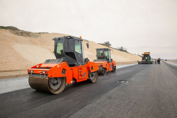 An attractive new orange road roller with a climate controlled enclosed cab sits on an unfinished new road.