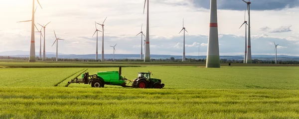 Foto op Plexiglas Schilderachtige landschapsmening grote moderne tractormachine met sproeiapparatuur die kunstmest sproeit in koolzaadlandbouwgebied tegen windmolen. Akkerbouw duurzame energie © Kirill Gorlov
