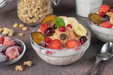 Granola crispy muesli with natural yogurt, frozen berries and fruit and nuts in glass bowls on brown background, healthy breakfast