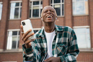Obraz na płótnie Canvas Authentic portrait of emotional hipster guy with stylish hairstyle using mobile phone listening music, laughing outdoors. Happy African American man holding smartphone communication on the street 