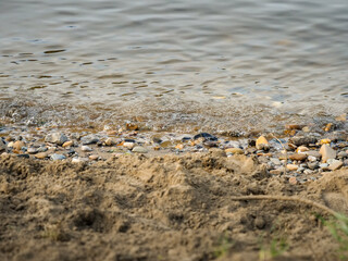 Water waves are moving to the shoreline of a river or a lake. Natural motion and fluidity. Wet stones and sand on the coast. Close-up of a beach in a natural environment.