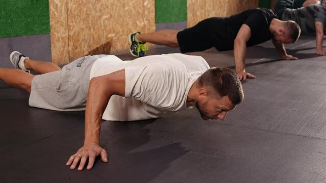 Full-length shot groups athletes engaged in sport, looking away while doing push-ups. Training, active lifestyle, team concept. Horizontal shot. Fit young people doing pushups in gym looking focused