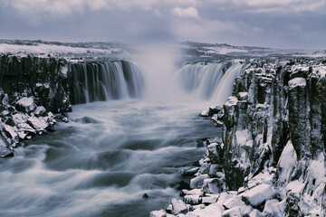 Centered purplish waterfall over dark basalt columns covered in snow, long exposure water stream with snow covered stone blocks in foreground, cloudy purplish sky