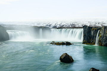Bright waterfall with stone cliff on right, greenish cold water with centered stone in foreground, background snow covered hill disappearing in white foggy sky