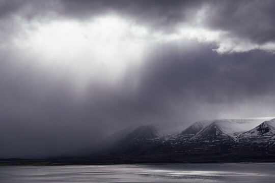 Sunlight breaking through dramatic purplish storm clouds, in background scandinavian snow covered mountain formation partly hidden by heavy rainfall, foreground dark lake reflecting light