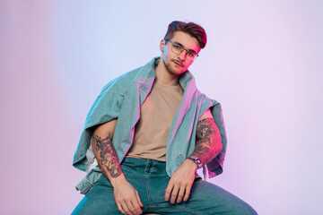 Fashionable handsome man hipster with hairstyle and vintage eyewear in trendy jeans outfit with jeans jacket with tattoos on arms sits in studio with creative pink blue lights