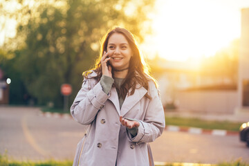 smiling beautiful girl with long hair in a grey trench coat using smartphone in the spring on the street in sunlight