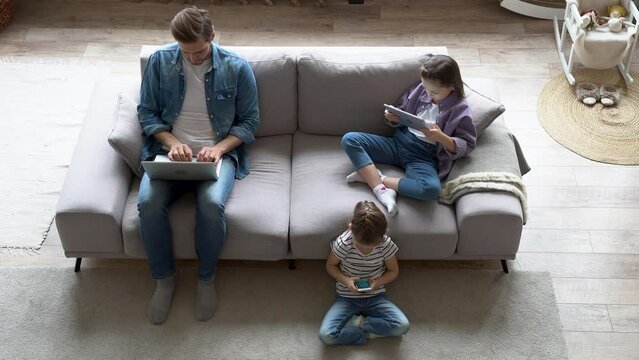 Addicted to gadgets father and kids busy on laptop, tablet and cellphone while sitting on sofa in living room. Modern family with children spending joint leisure time using gadgets