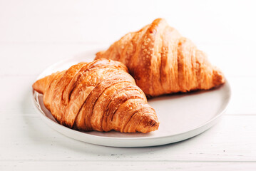Plate with two fresh croissants on white wooden table.