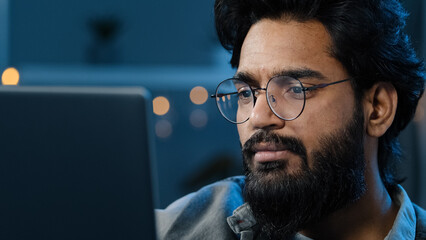 Thoughtful arabian indian bearded adult man thinking of problem solution working on laptop at home doubtful male face in glasses looking at computer screen watching video late night darkness overtime - obrazy, fototapety, plakaty