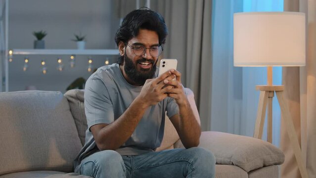Smiling Arabian Indian man bearded male looking in smartphone smile texting with friend girlfriend messaging chatting at home sitting on couch at night playing game in mobile app watching funny video