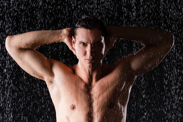 wet man posing with hands behind head under water drops on black background.