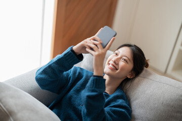 Image of smiling attractive asian woman using mobile phone while lying on sofa at home