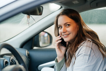 Fototapeta na wymiar beautiful girl with long hair in a grey trench coat using smartphone call gets into the car