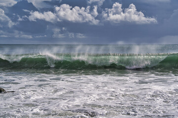 A Turquoise wave breaking on a beach in sea with silver clouds