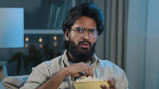 Interested Attentive Arabic Hispanic Indian Bearded Man Guy In Glasses Eating Popcorn Watching TV Show Series Horror Movie Late Night Feeling Scary Shocked Scared Reaction Disgust Horrible Thriller