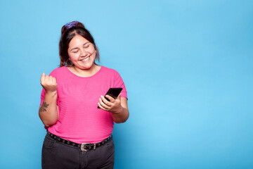 Young curvy latina woman, celebrating good news with fist raised, holding mobile phone. Indoor...