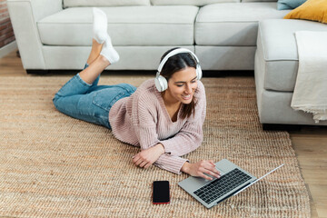 Pretty young woman listening music with headphone while working with laptop lying on the floor at...