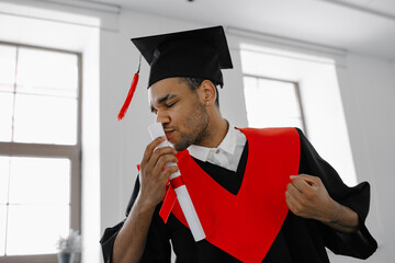 A Black student in graduate gown and square cap who is happy to finish his studies shows his the...