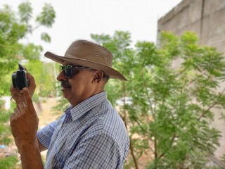 Picture of a person wearing sunglasses and hat out in a sanctuary in summers with green leaves in background shooting pictures with his camera

