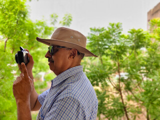 Picture of a person wearing sunglasses and hat out in a sanctuary in summers with green leaves in background shooting pictures with his camera
