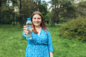 Beautiful young woman holding bottle against green nature background. Optimistic redhead girl walking in city summer park. Closeup female hand holding water at green park outdoor. Healthy concept.