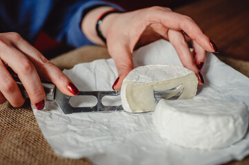 Obraz na płótnie Canvas Cut Camembert cheese, French soft cheese with white mold in the hands of a woman on the background of a craft textile tablecloth, close-up. Knife for slicing cheese. Cheesemaker cuts cheese.