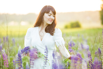 Beautiful woman in white sundress enjoying the summer nature. Picking colourful flowers, breathing fresh air and floral scent, walking in the sunny field of lupins. Happiness connect