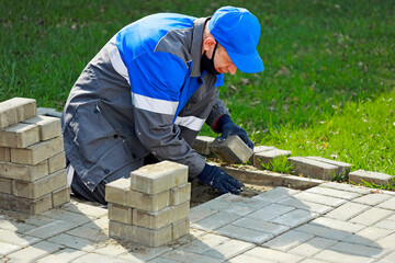 Bricklayer in work clothes sits on sidewalk and lays out paving slabs. Sight of working man in open air. Professional builder makes arrangement of territory summer day. Real scene.