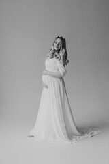 Fototapeta na wymiar Blonde pregnant woman in a white dress with a wreath on her head in the studio on a white background holds her hand on her stomach black and white