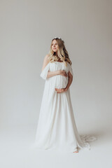 Fototapeta na wymiar Blonde pregnant woman in a white dress with a wreath on her head in the studio on a white background holds her hand on her stomach