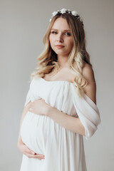 Blonde pregnant woman in a white dress with a wreath on her head in the studio on a white background holds her hand on her stomach