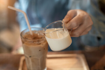 A hand pouring a glass of milk cream into iced latte coffee on a wooden bar over a cafe glass window reflex at a Cafe coffee shop. Cold brew refreshment summer drink with copy space. Selective focus