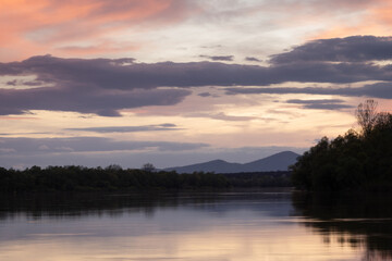 Landscape of river and mountain silhouette at dusk, Sava river with forested shore and Motajica mountain with clouds in sky and orange glow in clouds during dusk