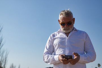 Mature man, gray haired, bearded, sunglasses, white shirt, checking social networks on his cell...