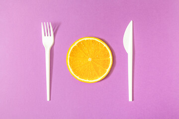 Minimal composition with orange slice, fork and knife on bright purple background. Healthy meal...