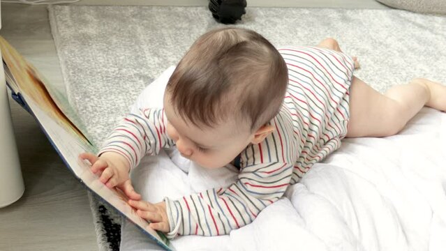 adorable cute little baby boy is lying on floor with a open book in front. toddler is playing with hands on pages surfaces. 4k video. education concept. story telling