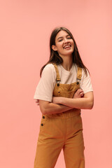 Cool young caucasian girl smiles looking into camera with arms crossed standing in pink background. Teenager with dark, straight hair wears T-shirt and jumpsuit. Lifestyle, leisure concept. 