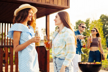 Two girls are drinking drinks, in the background a group of friends having fun grilling meat enjoying barbecue