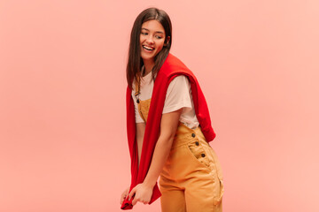 Cheerful young caucasian brunette girl with straight hair looks away posing on pink background. Lady wears white T-shirt, yellow jumpsuit and red sweatshirt on top.