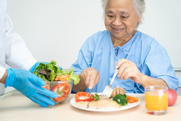 Asian senior or elderly old lady woman patient eating Salmon steak breakfast with vegetable healthy food while sitting and hungry on bed in hospital.