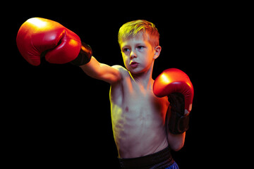 Little sportive boy, beginner boxer in red boxer gloves and shorts training isolated on dark...
